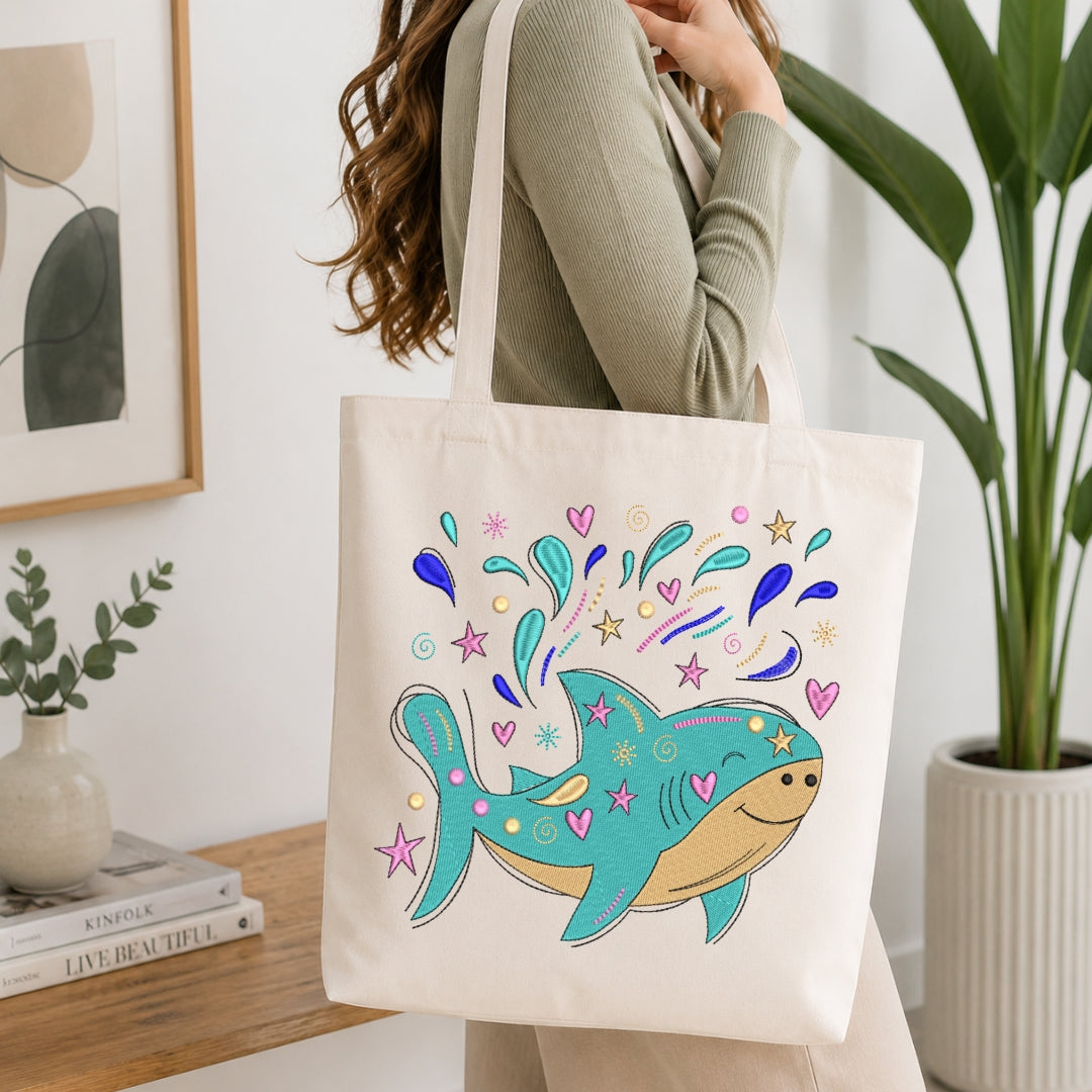 A woman wearing a sage green ribbed long-sleeve top carries a cream canvas tote bag with a colorful cartoon shark graphic. The background shows a bright, modern room with a wooden table, books, a potted plant, and framed wall art.