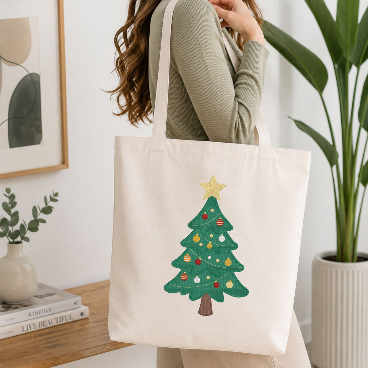 Woman carrying a white natural canvas tote bag featuring a classic decorated Christmas tree machine embroidery design in a bright, modern interior setting
