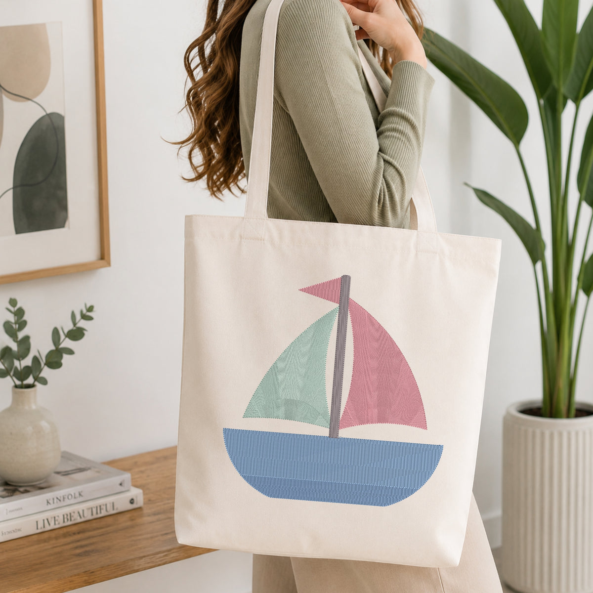A person holding a natural canvas tote bag printed with an embroidered colorful sailboat design, set against a bright interior room background with green plants.