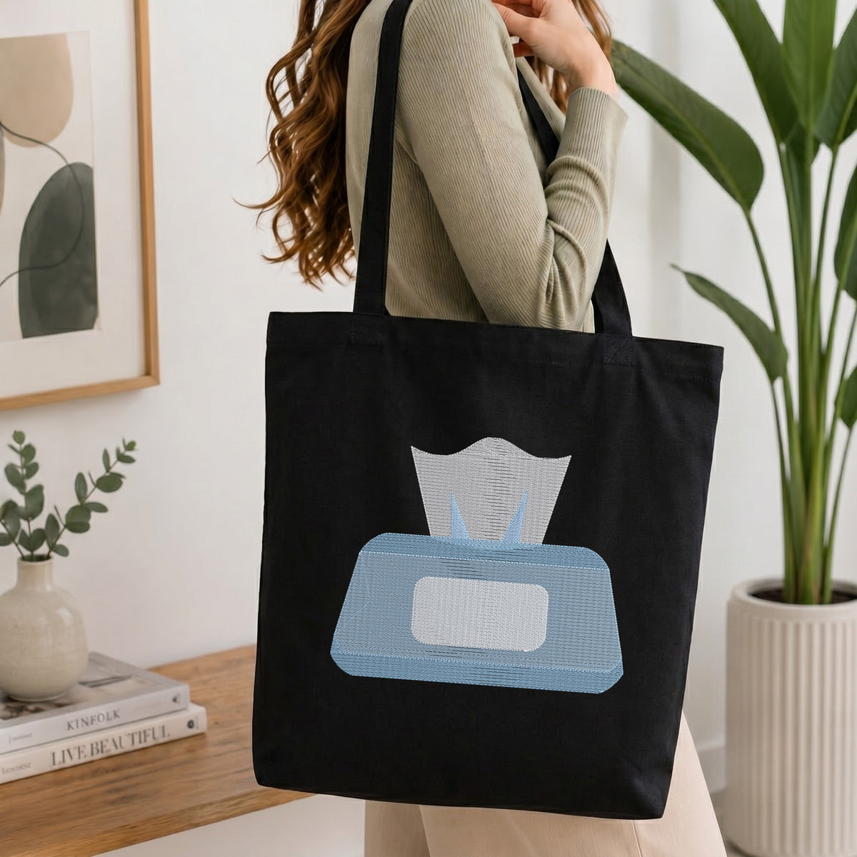 A woman wearing a beige ribbed sweater carries a black canvas tote bag with a light blue and white tissue box graphic on the front, set in a bright, modern room with a wooden table, framed art, and a potted plant.