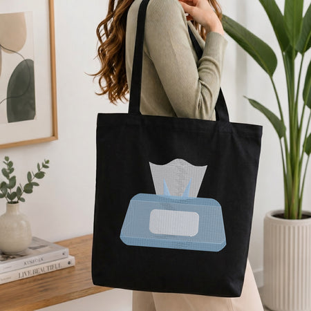 A woman wearing a beige ribbed sweater carries a black canvas tote bag with a light blue and white tissue box graphic on the front, set in a bright, modern room with a wooden table, framed art, and a potted plant.