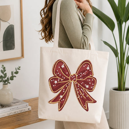 A woman wearing a sage green ribbed sweater carries a cream canvas tote bag with a large embroidered red bow design with gold and white star details, set against a bright, modern interior background with a potted plant and books.