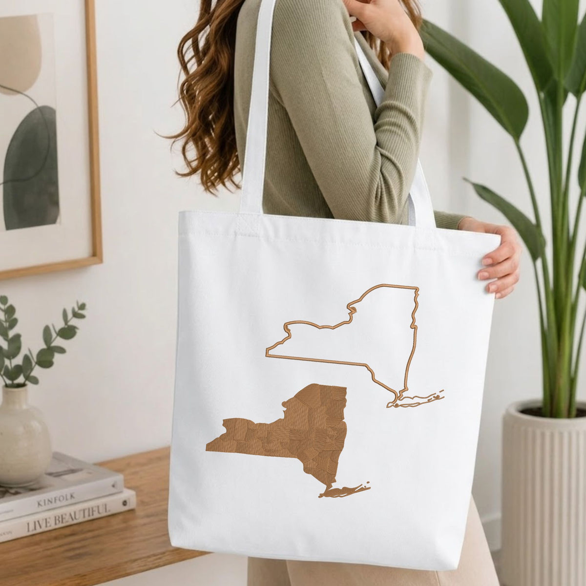  Woman carrying a white natural canvas tote bag featuring an embroidered New York state map design in a bright, modern interior setting.   