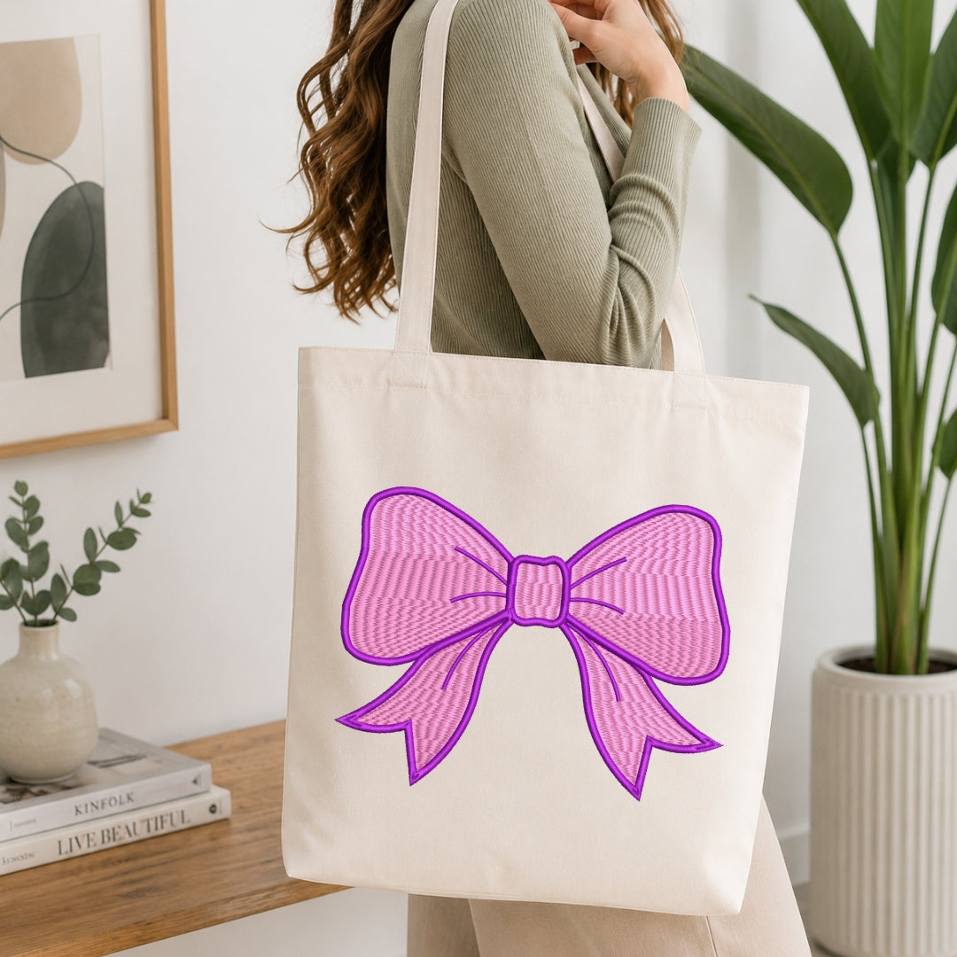 A person wearing a ribbed sage green long-sleeve top carries a cream canvas tote bag with a large pink and purple bow graphic on the front, set in a bright, modern room with a wooden table and potted plants.