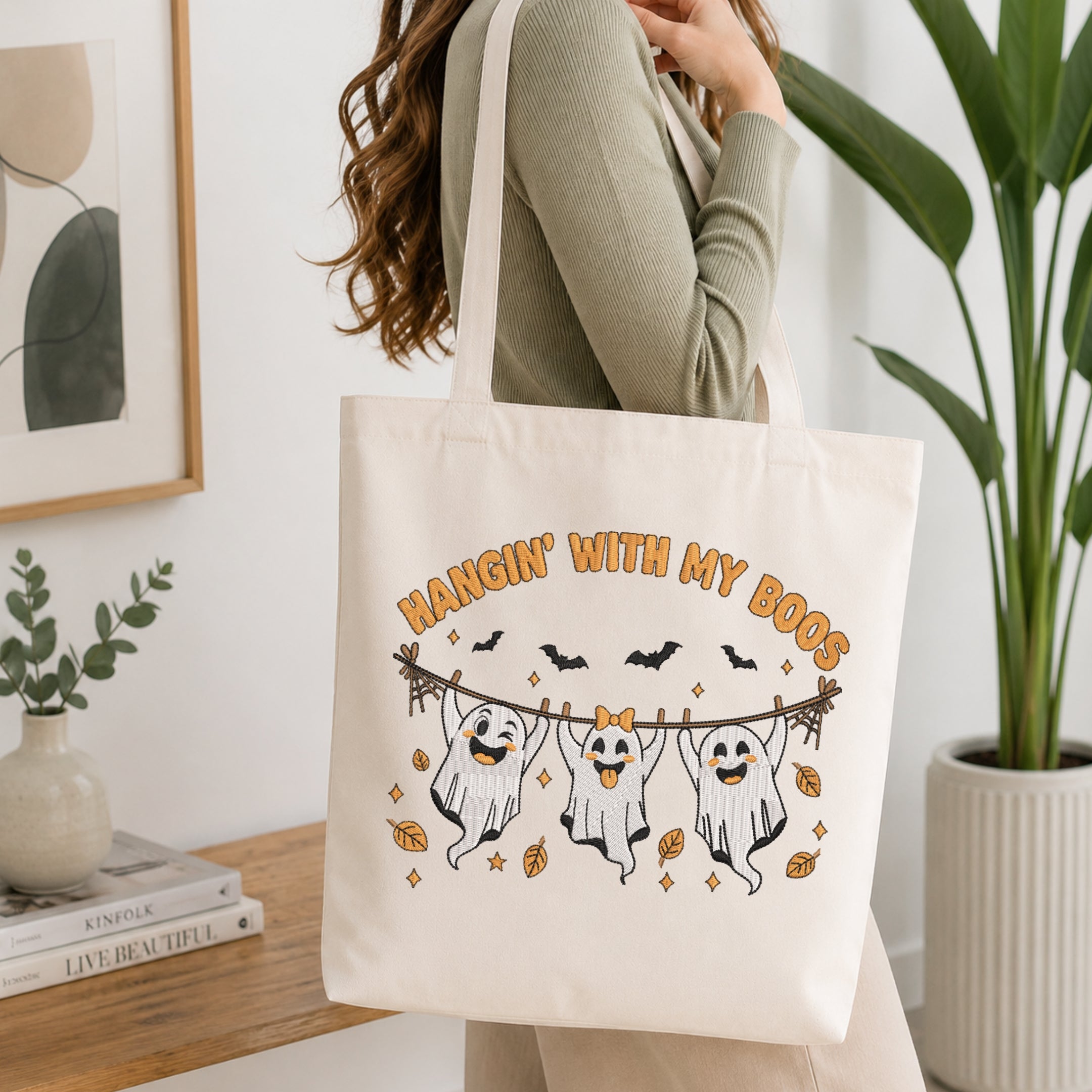 A woman wearing a sage green ribbed long-sleeve top carries a cream canvas tote bag with a Halloween graphic that reads 'HANGIN' WITH MY BOOS' and shows three ghosts hanging from a wire with bats and leaves. The photo is taken in a bright, modern room with a wooden shelf, framed art, and a potted plant.
