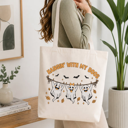 A woman wearing a sage green ribbed long-sleeve top carries a cream canvas tote bag with a Halloween graphic that reads 'HANGIN' WITH MY BOOS' and shows three ghosts hanging from a wire with bats and leaves. The photo is taken in a bright, modern room with a wooden shelf, framed art, and a potted plant.