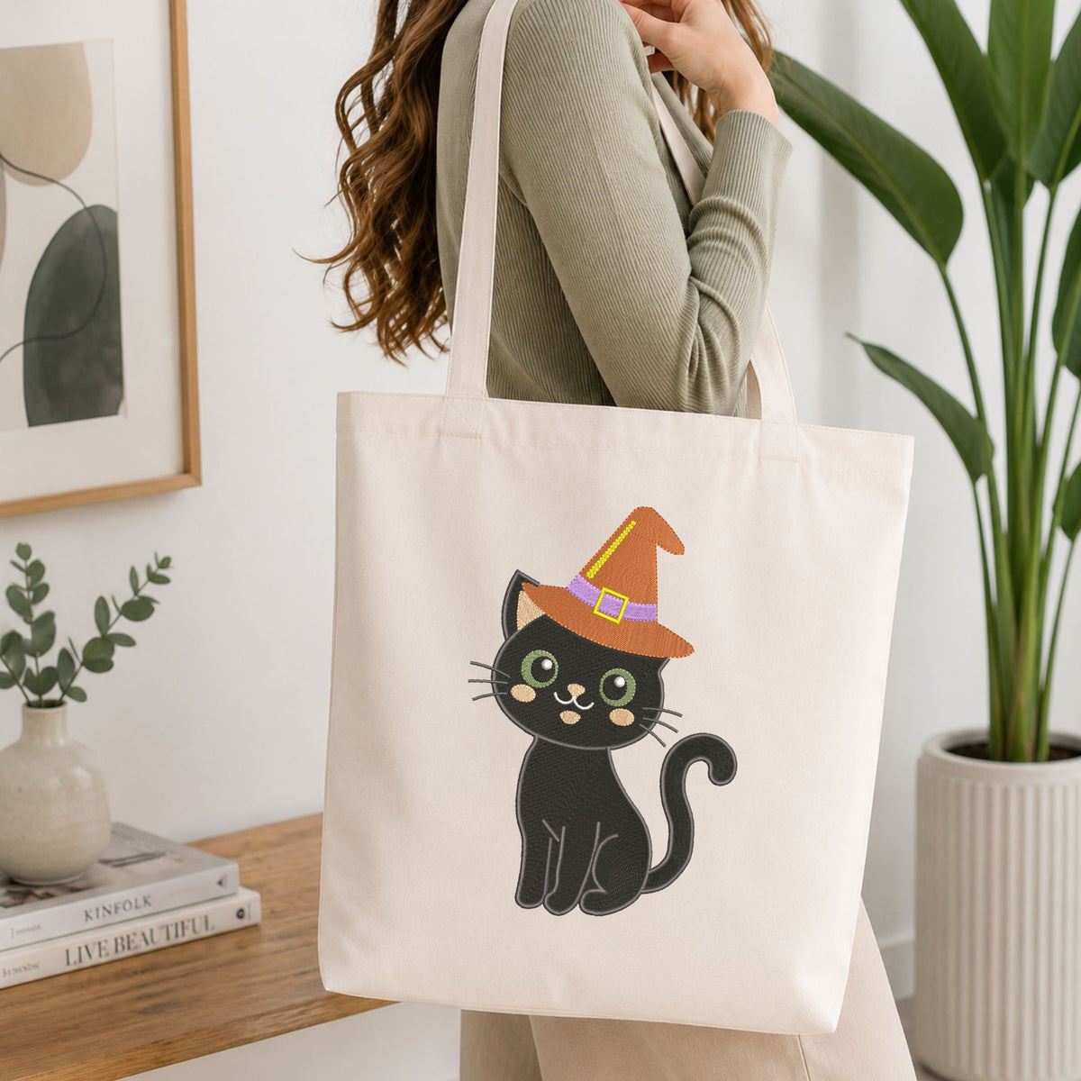 Woman carrying a white natural canvas tote bag featuring an embroidered black cat in a witch hat design in a bright, modern interior setting.