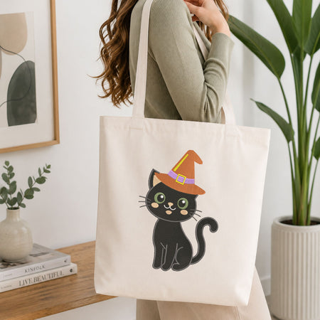 Woman carrying a white natural canvas tote bag featuring an embroidered black cat in a witch hat design in a bright, modern interior setting.