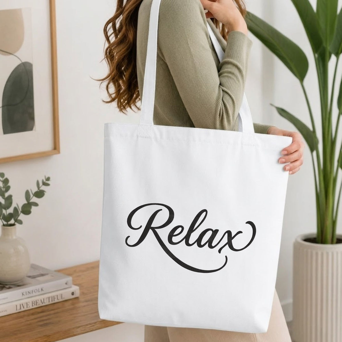 Woman carrying a white natural canvas tote bag featuring an embroidered "Relax" script design in a bright, modern interior setting