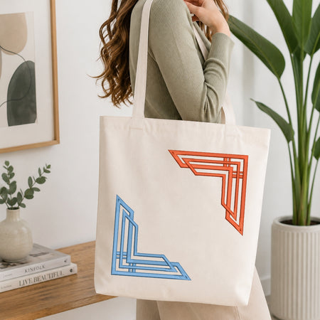 A woman wearing a sage green ribbed sweater carries a cream canvas tote bag with geometric orange and blue line art corner designs, set in a bright, modern room with a wooden table, books, and a potted plant.