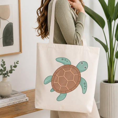 A woman wearing a sage green ribbed sweater carries a cream canvas tote bag with a large embroidered sea turtle design. The background is a bright, modern room with a wooden shelf, framed art, a small plant, and a large green potted plant.