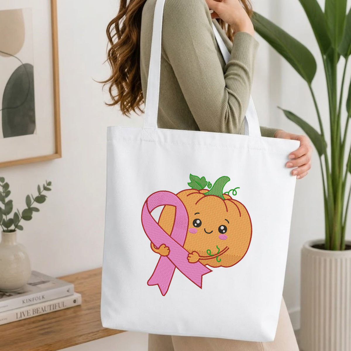 Woman carrying a white natural canvas tote bag featuring an embroidered kawaii pumpkin hugging a pink ribbon design in a bright, modern interior setting.