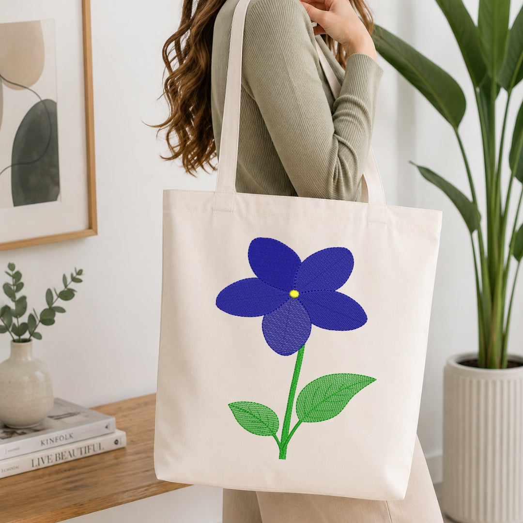 A woman carries a cream canvas tote bag featuring a large embroidered blue flower with green leaves and a small yellow center, shown in a bright, modern room with plants and a wooden table.