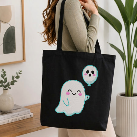 A person wearing a beige ribbed sweater carries a black canvas tote bag with a cute cartoon ghost and balloon graphic. The setting is a bright, modern room with a wooden table, books, a small plant, and a large potted plant in the background.