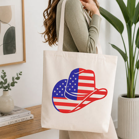 A woman wearing a sage green ribbed sweater carries a cream canvas tote bag with a large graphic of a cowboy hat filled with the American flag pattern, set in a bright, modern room with a wooden table and potted plants.