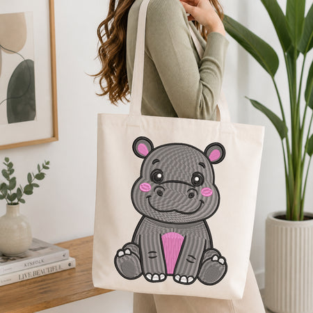 A woman wearing a sage green ribbed long-sleeve top carries a cream canvas tote bag with a cute cartoon hippo design. The hippo is gray with pink inner ears, pink cheeks, and a pink belly patch.