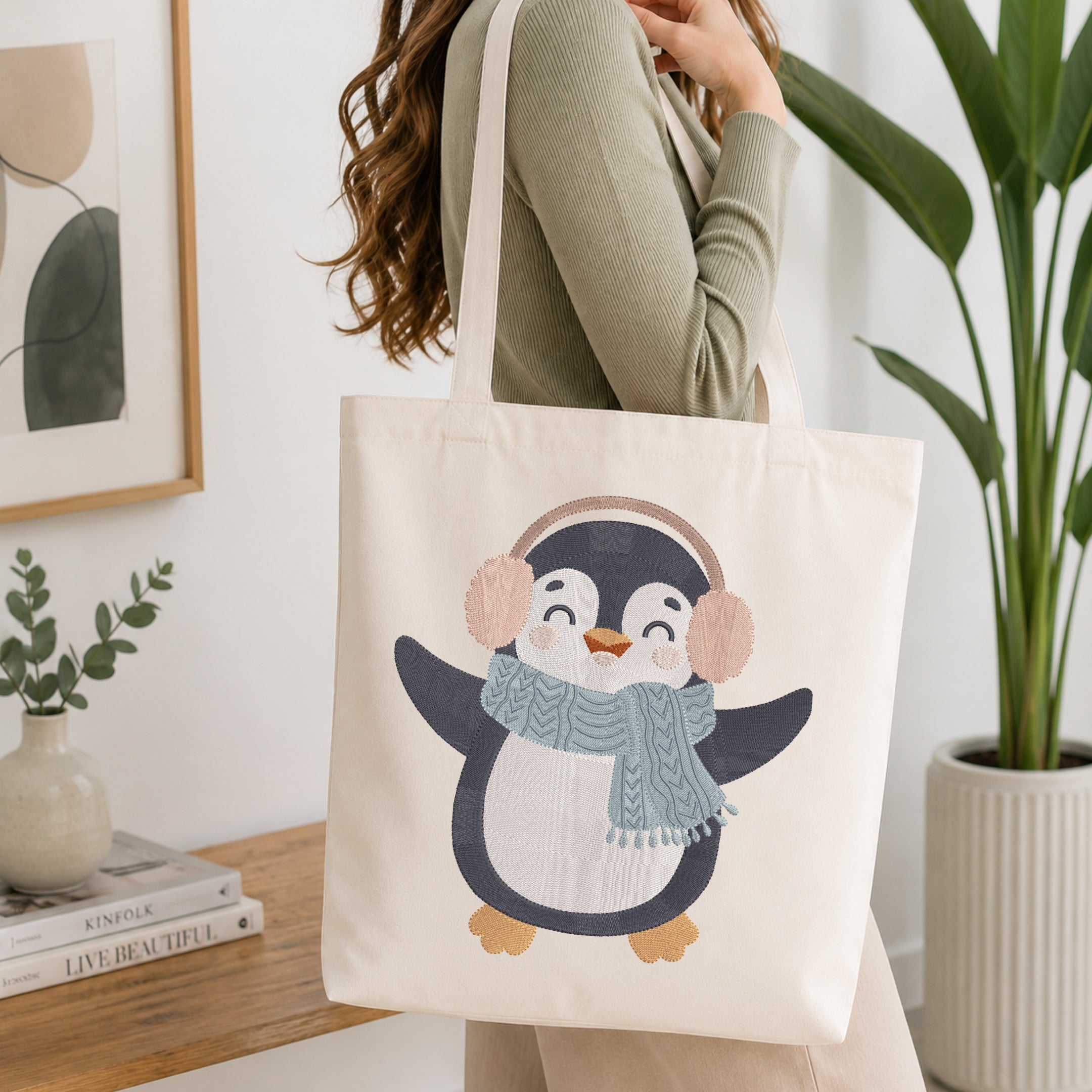 A woman wearing a sage green ribbed sweater carries a cream canvas tote bag with a cute penguin illustration. The penguin is wearing a gray knit scarf and pink earmuffs.