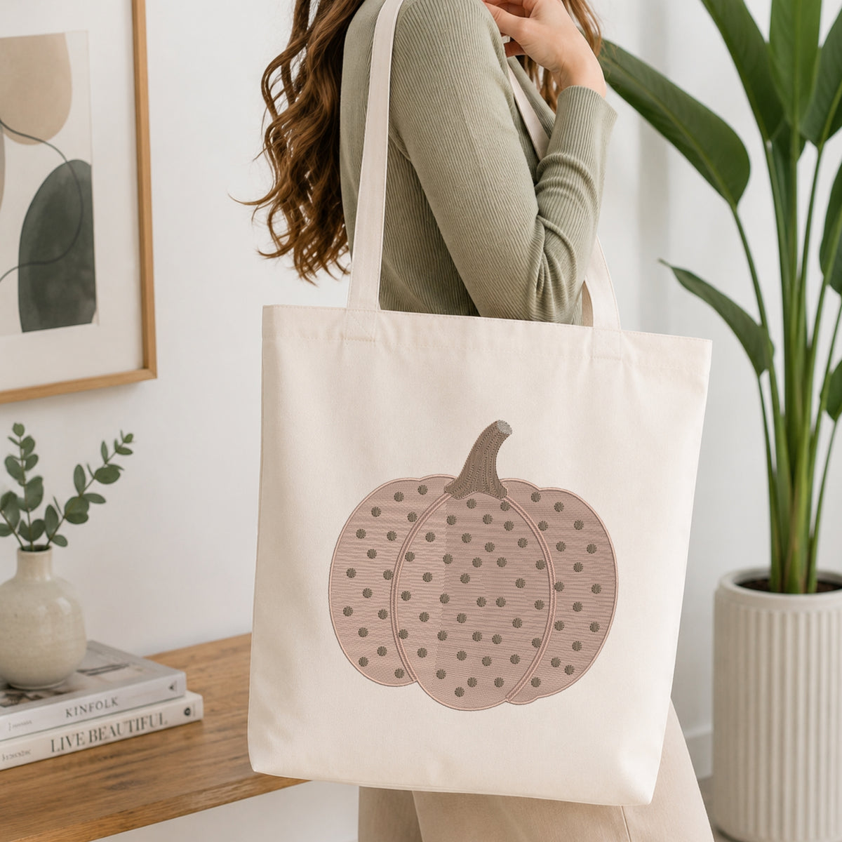 A woman wearing a sage green ribbed long-sleeve top carries a cream canvas tote bag with a large dusty pink pumpkin graphic featuring dark polka dots, set against a bright, modern interior background with a wooden table, books, and a potted plant.