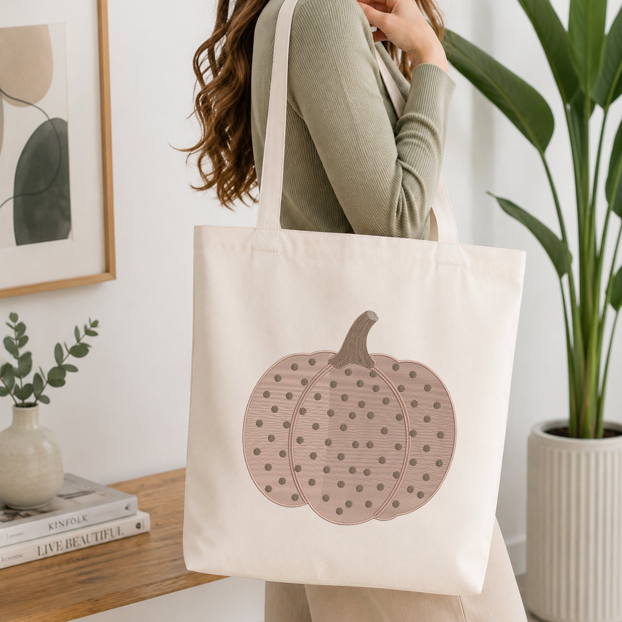 A woman wearing a sage green ribbed long-sleeve top carries a cream canvas tote bag with a large dusty pink pumpkin graphic featuring dark polka dots, set against a bright, modern interior background with a wooden table, books, and a potted plant.