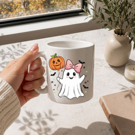 A hand holding a white ceramic mug with a cute Halloween graphic of a white ghost wearing a pink bow next to an orange jack-o'-lantern pumpkin, set on a light marble tabletop near a window.