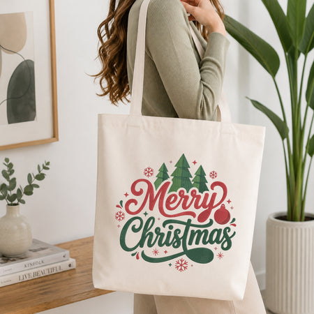 A woman wearing a sage green ribbed sweater carries a cream canvas tote bag with a 'Merry Christmas' graphic featuring red and green lettering, pine trees, and snowflakes in a bright, modern room.