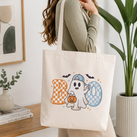 A woman wearing a sage green ribbed long-sleeve top carries a cream canvas tote bag with a colorful 'BOO' Halloween graphic featuring a cartoon ghost, pumpkins, bats, and checkered letters. The background is a bright, modern room with a wooden shelf, framed art, and a potted plant.