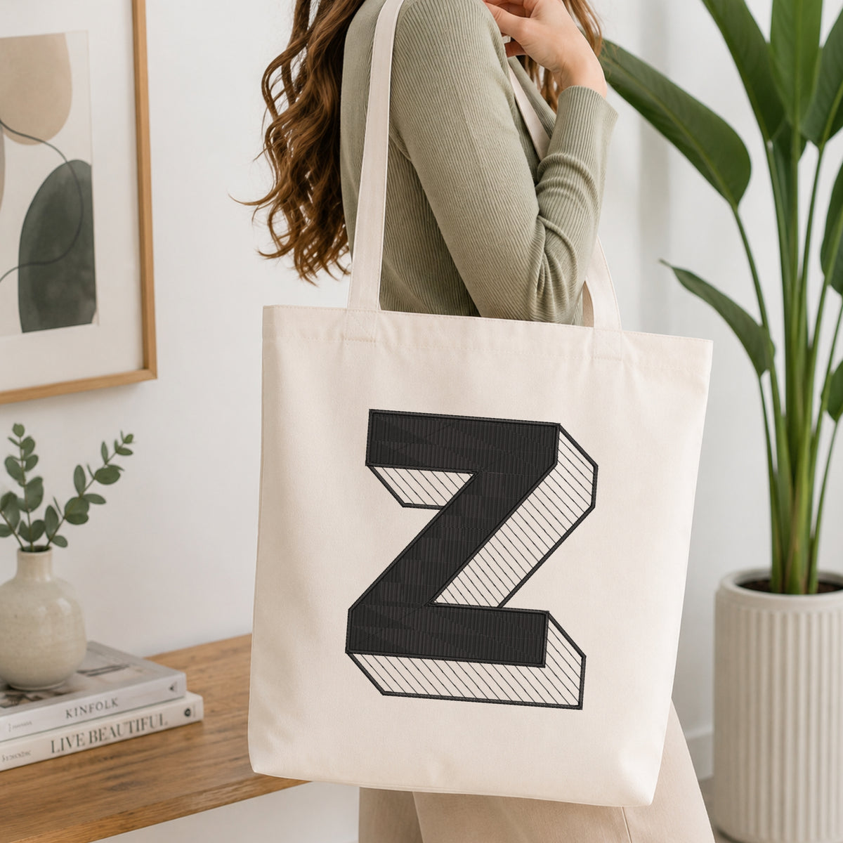 A woman wearing a sage green ribbed long-sleeve top carries a cream canvas tote bag with a large black 3D-effect letter 'Z' printed on the front, set in a bright, modern room with a wooden console table and a potted plant.