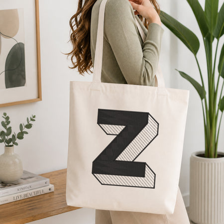 A woman wearing a sage green ribbed long-sleeve top carries a cream canvas tote bag with a large black 3D-effect letter 'Z' printed on the front, set in a bright, modern room with a wooden console table and a potted plant.