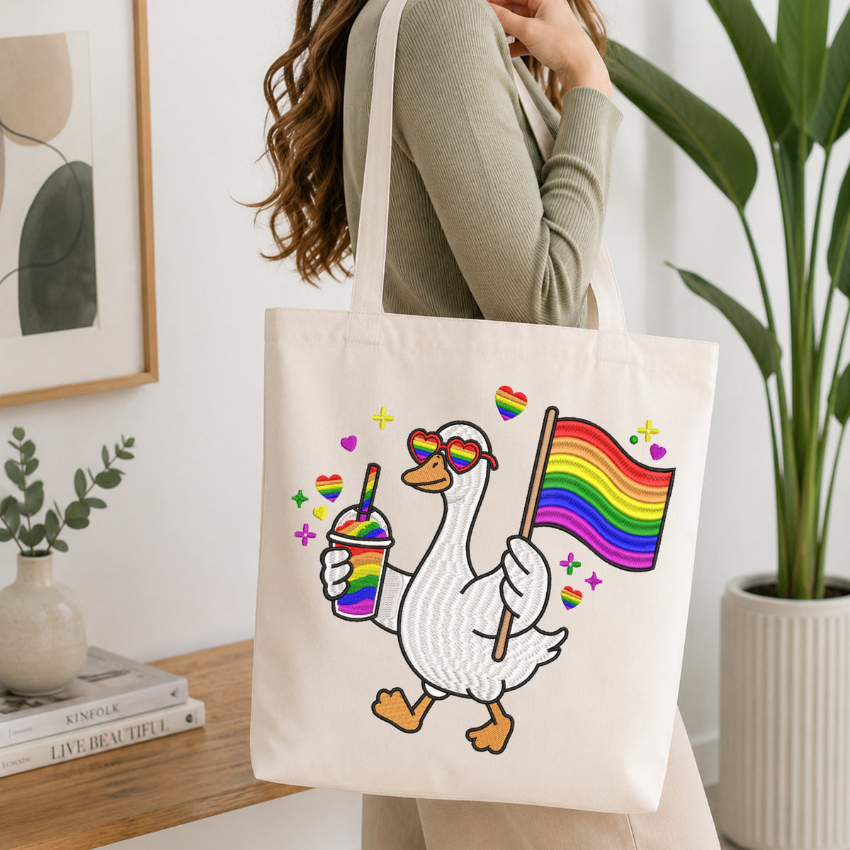 A woman carries a cream canvas tote bag featuring a cartoon white duck wearing rainbow heart sunglasses and holding a rainbow pride flag, with small rainbow drink and heart accents. The scene is set in a bright, modern room with a wooden table, books, and a potted plant.