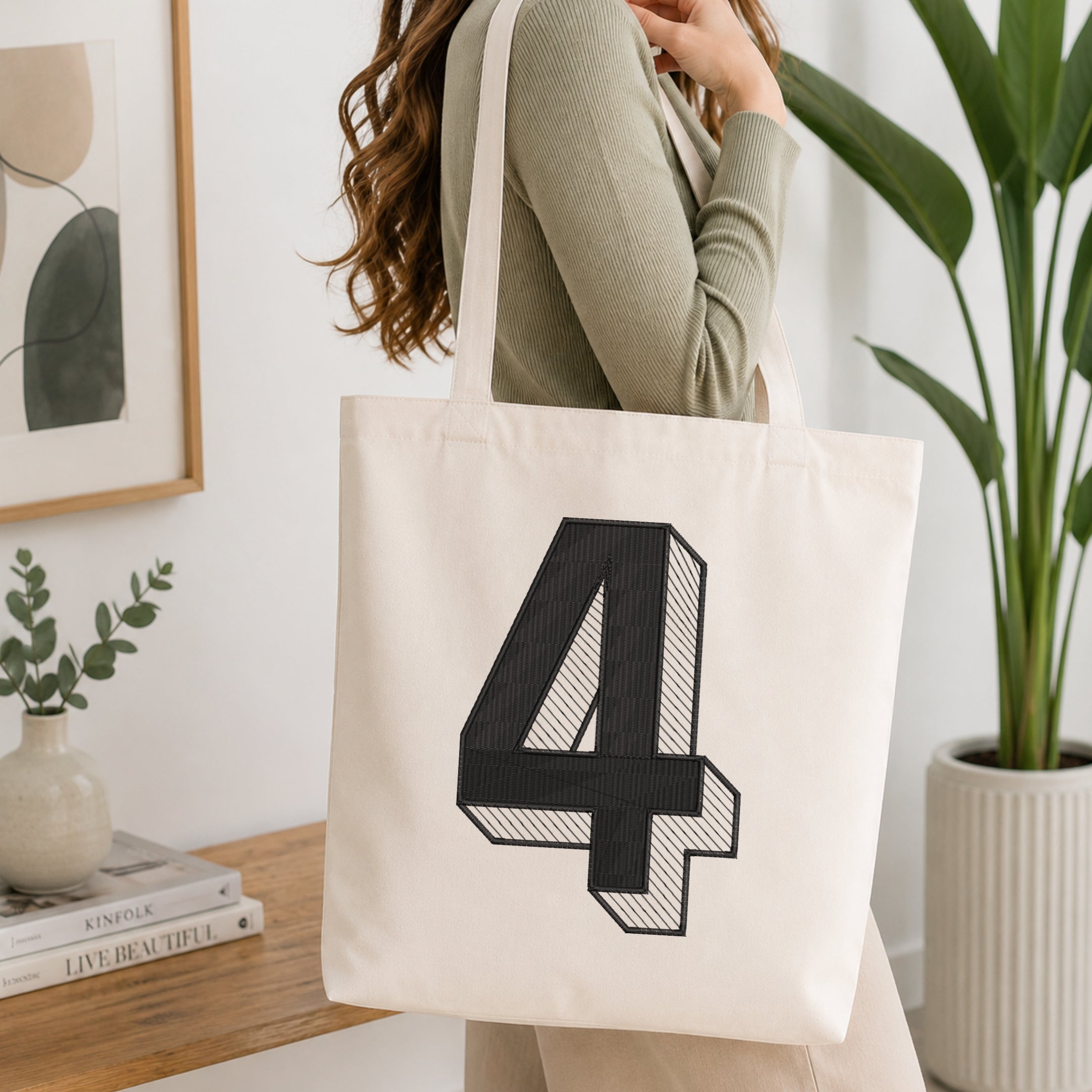 A woman wearing a sage green ribbed long-sleeve top carries a cream canvas tote bag with a large black 3D-style number 4 printed on the front, set in a bright, modern room with a wooden table, framed art, and a potted plant.