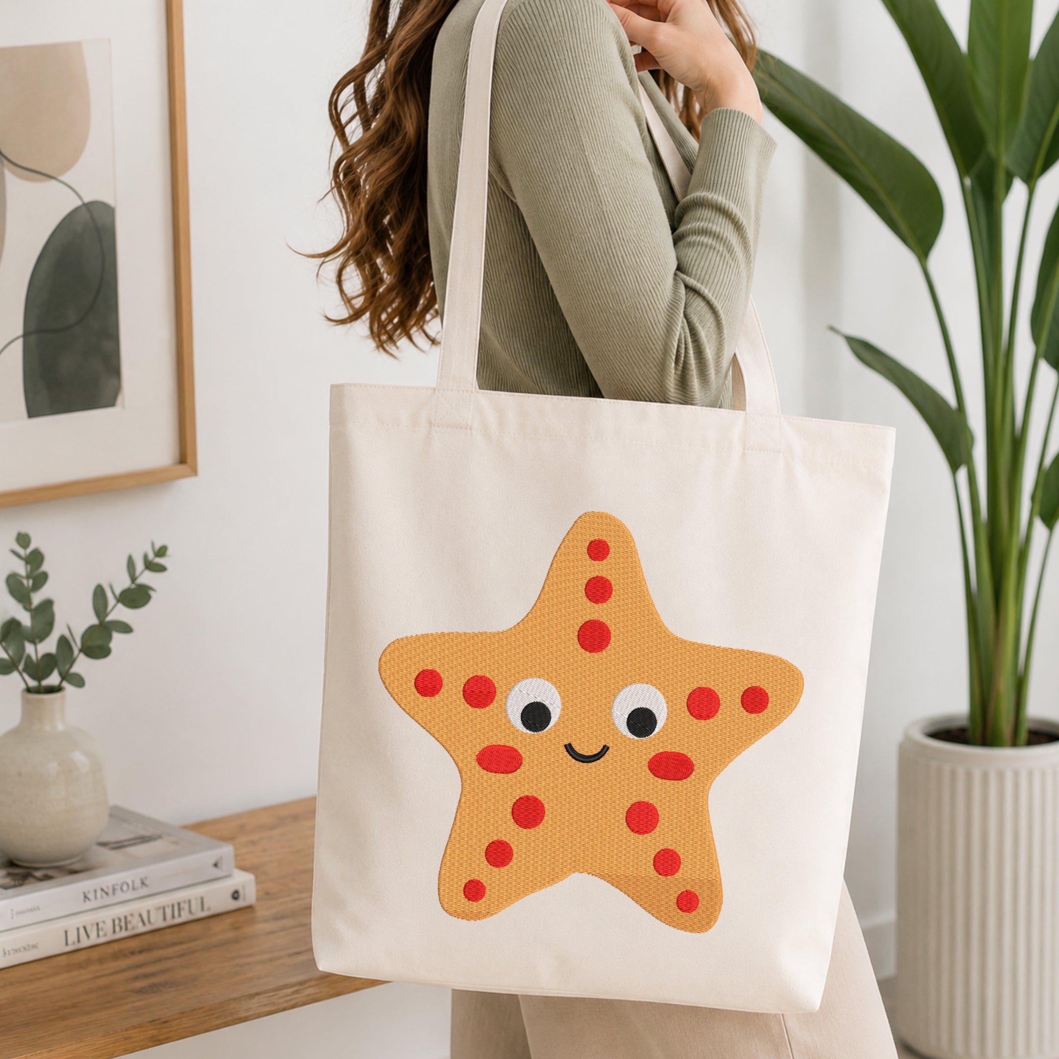 A woman wearing a sage green ribbed sweater carries a cream canvas tote bag with a large orange starfish applique featuring red dots and a smiling face, set in a bright, modern room with a wooden table and potted plants.