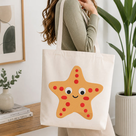 A woman wearing a sage green ribbed sweater carries a cream canvas tote bag with a large orange starfish applique featuring red dots and a smiling face, set in a bright, modern room with a wooden table and potted plants.