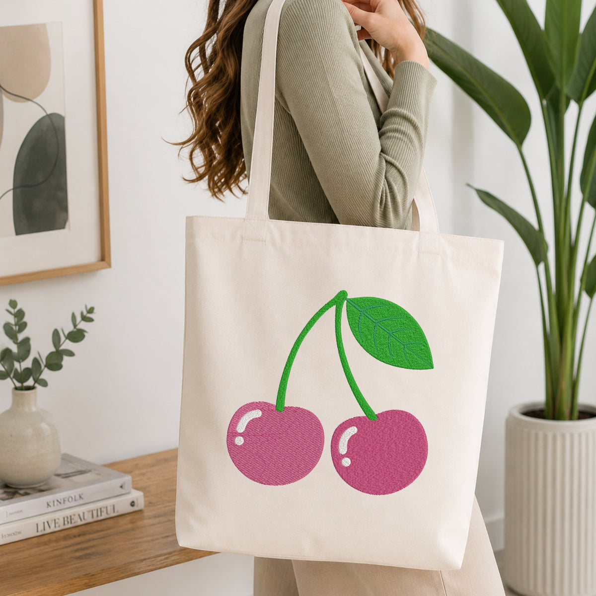 A woman wearing a sage green ribbed sweater carries a cream canvas tote bag featuring a large pink and green cherry graphic on the front, set in a bright, modern room with a wooden table and potted plants.