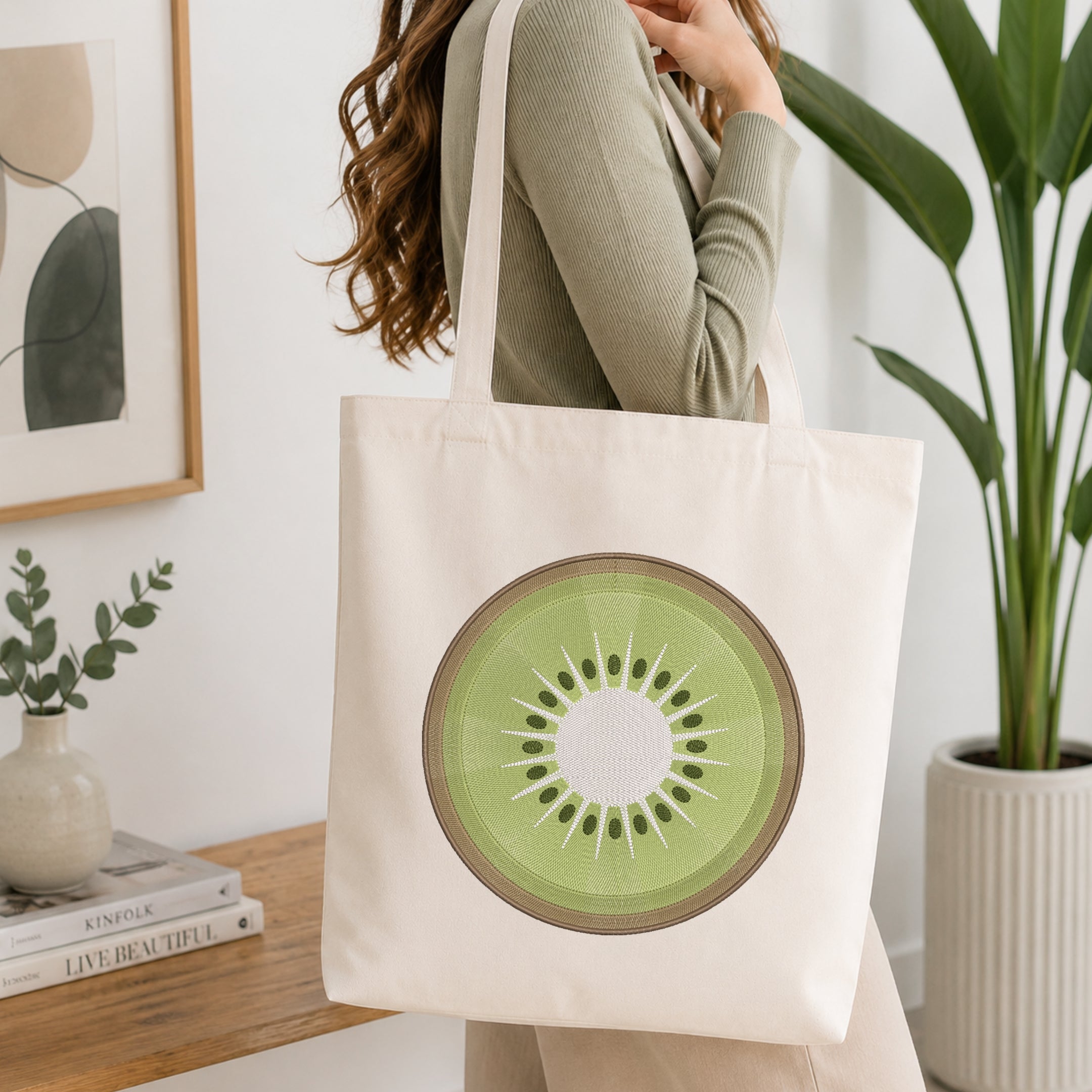 A woman wearing a sage green ribbed sweater carries a cream canvas tote bag featuring a large green kiwi slice graphic on the front, set against a bright, modern interior background with a wooden table, framed art, and a potted plant.