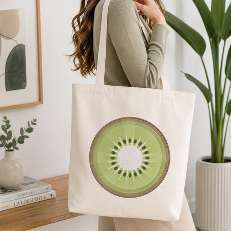 A woman wearing a sage green ribbed sweater carries a cream canvas tote bag featuring a large green kiwi slice graphic on the front, set against a bright, modern interior background with a wooden table, framed art, and a potted plant.