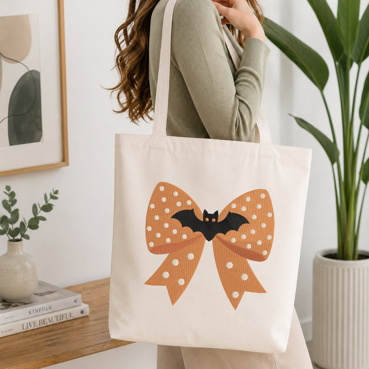 A person wearing a green long-sleeve top and carrying a natural canvas tote bag printed with the orange bat bow machine embroidery graphic.