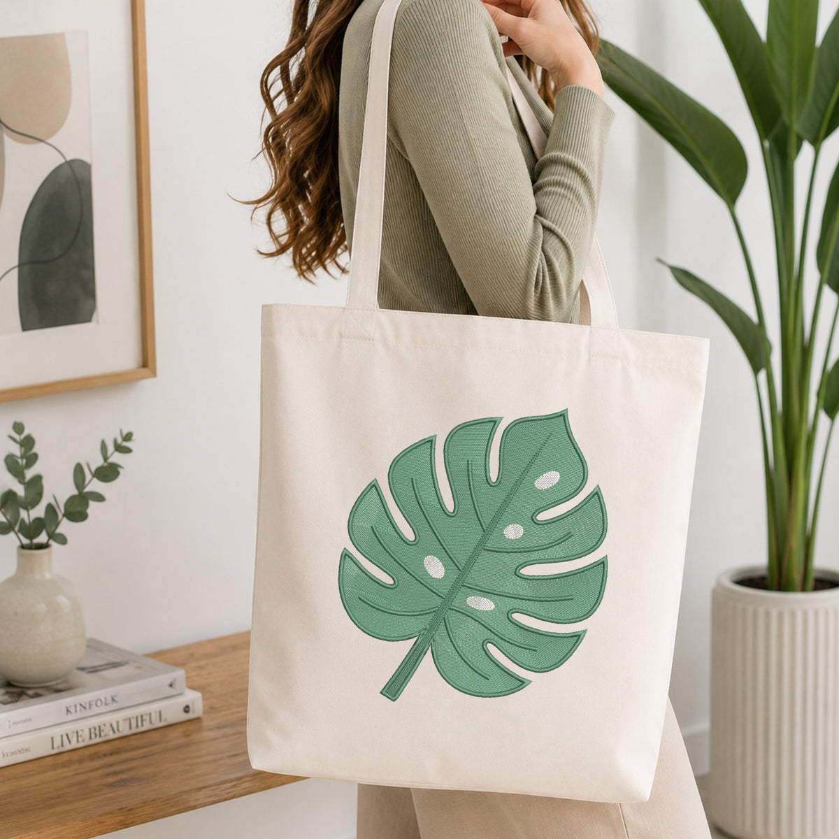 A woman wearing a sage green ribbed long-sleeve top carries a cream canvas tote bag with a large green monstera leaf print. The scene is set in a bright, modern room with a wooden shelf, a small potted plant, and a large green plant in the background.