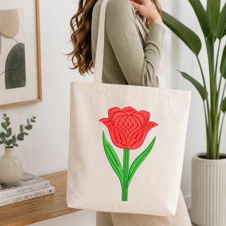 A woman wearing a sage green ribbed sweater carries a cream canvas tote bag with a large red tulip and green leaves printed on the front, set in a bright, modern room with a wooden table and potted plants.