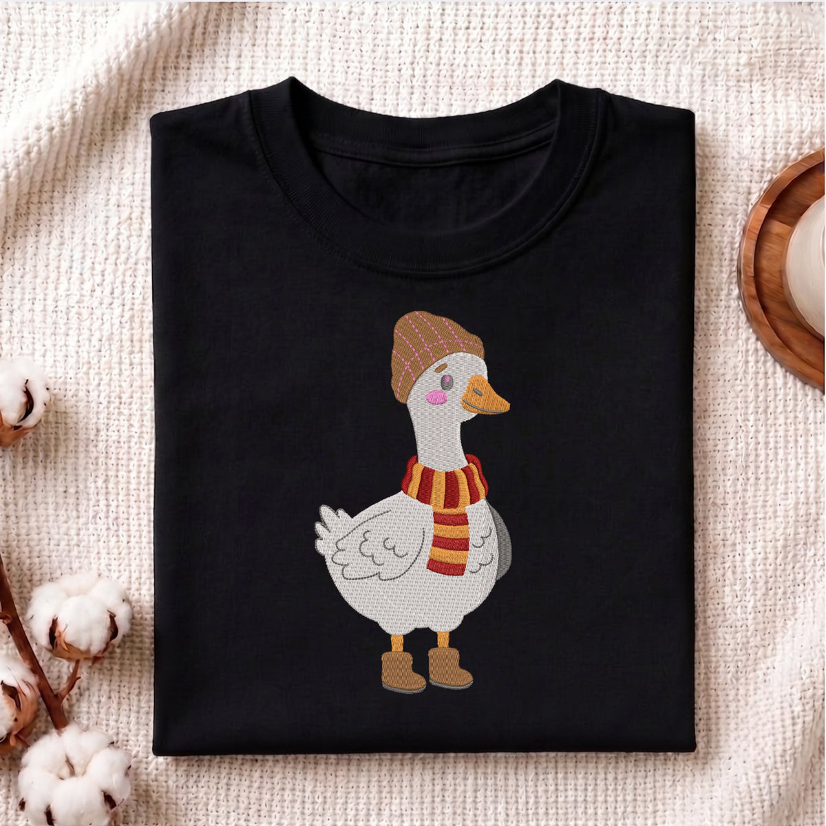 A folded black crewneck t-shirt featuring a cartoon illustration of a white goose wearing a brown plaid beanie and a red and yellow striped scarf, styled on a beige textured fabric background with cotton bolls.