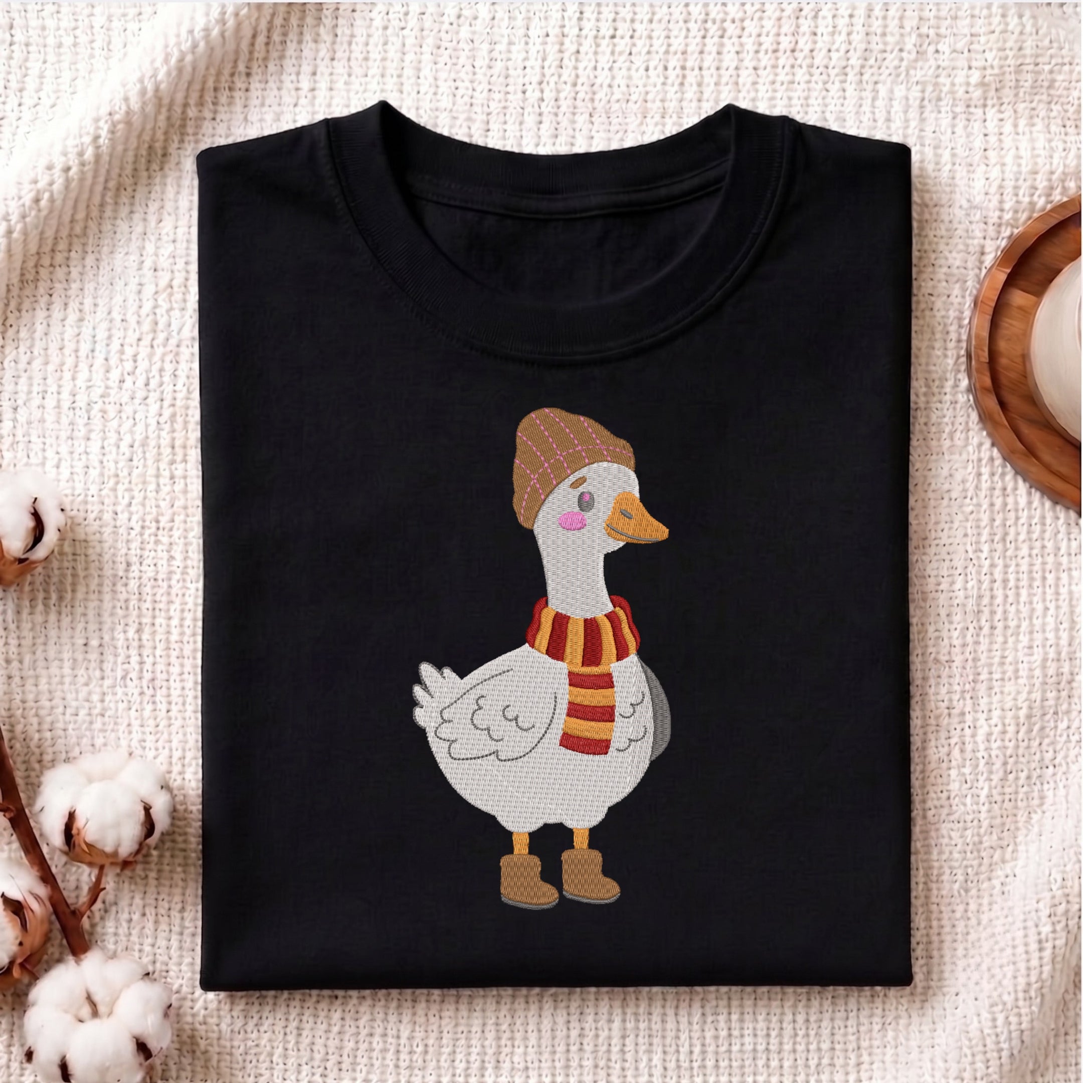 A folded black crewneck t-shirt featuring a cartoon illustration of a white goose wearing a brown plaid beanie and a red and yellow striped scarf, styled on a beige textured fabric background with cotton bolls.