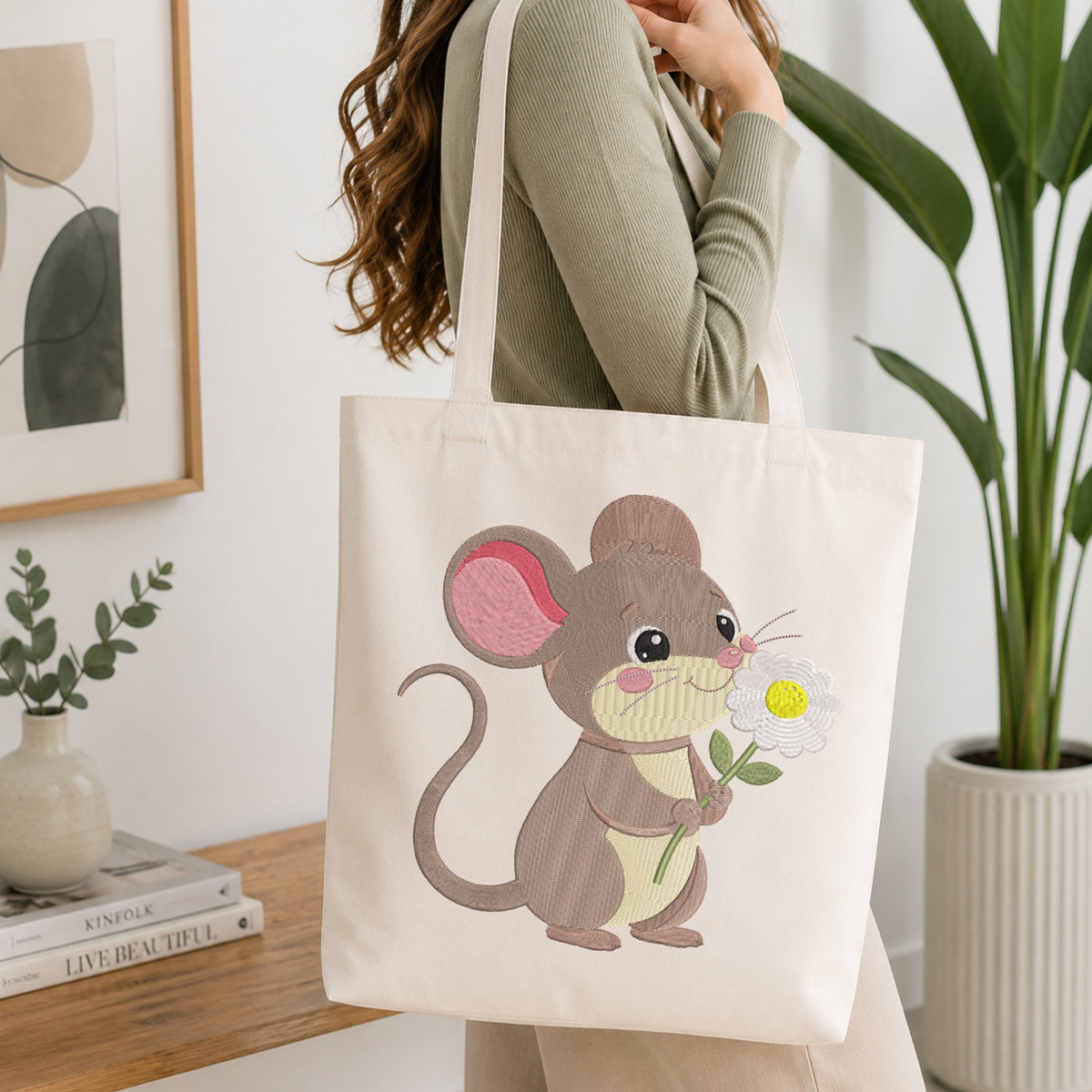 A woman wearing a sage green ribbed sweater carries a cream canvas tote bag with a cute embroidered mouse holding a daisy. The scene is set in a bright, modern room with a wooden table, books, a small plant, and a large green potted plant in the background.