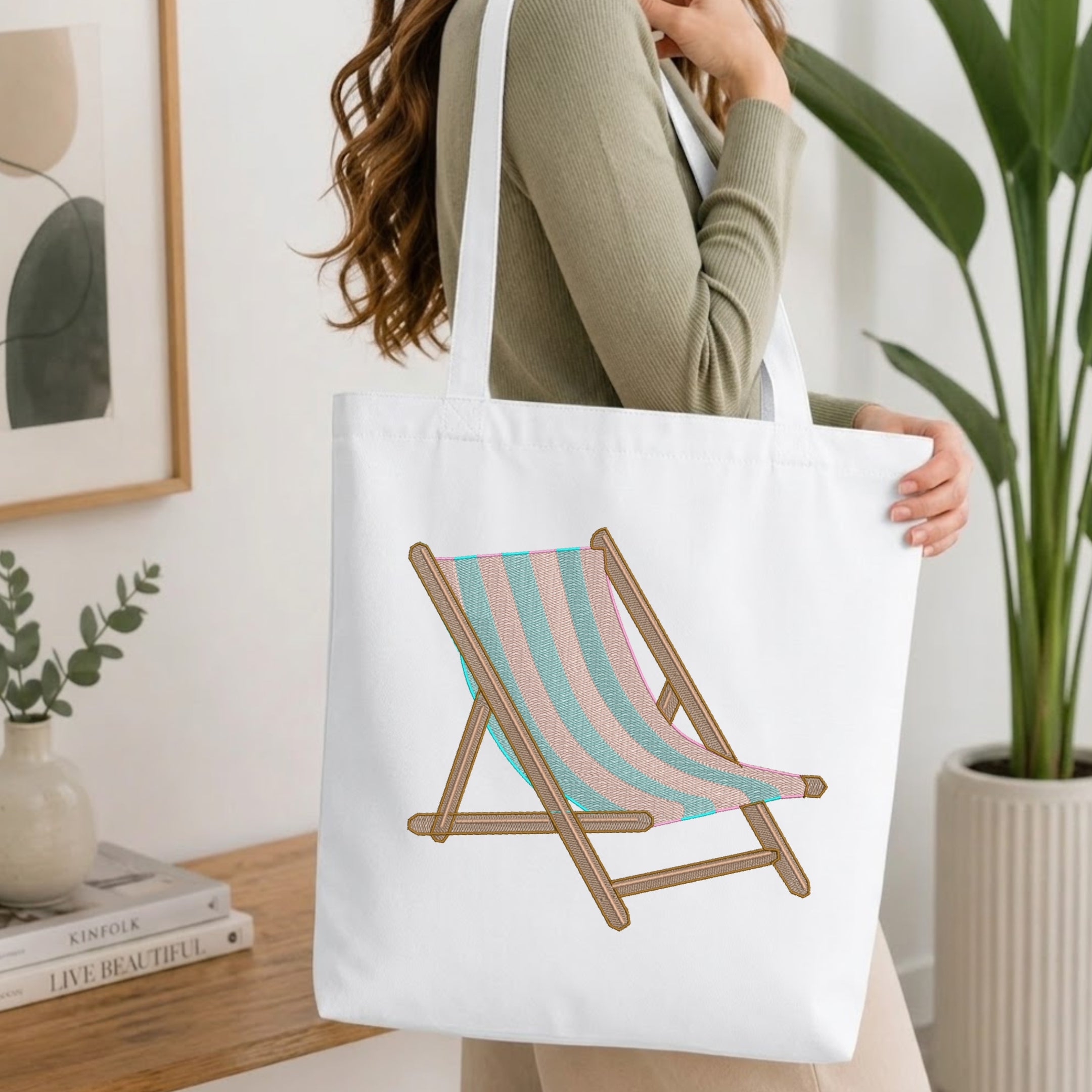 A woman wearing a sage green ribbed long-sleeve top carries a white canvas tote bag. The bag features a printed graphic of a wooden deck chair with pastel pink and light blue striped fabric.