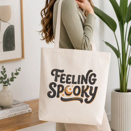 Woman carrying a white natural canvas tote bag featuring an embroidered "Feeling Spooky" design in a bright, modern interior setting.