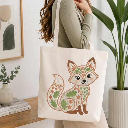 A person wearing a sage green ribbed sweater carries a cream canvas tote bag featuring a whimsical embroidered-style fox design in green, brown, and tan. The scene is set in a bright, modern room with a wooden table, books, a framed print, and a potted plant.