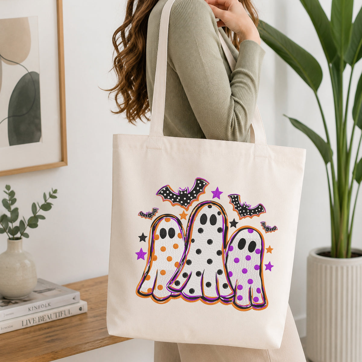 Woman carrying a white natural canvas tote bag featuring a cute Halloween ghosts and bats machine embroidery design in a bright, modern interior setting   