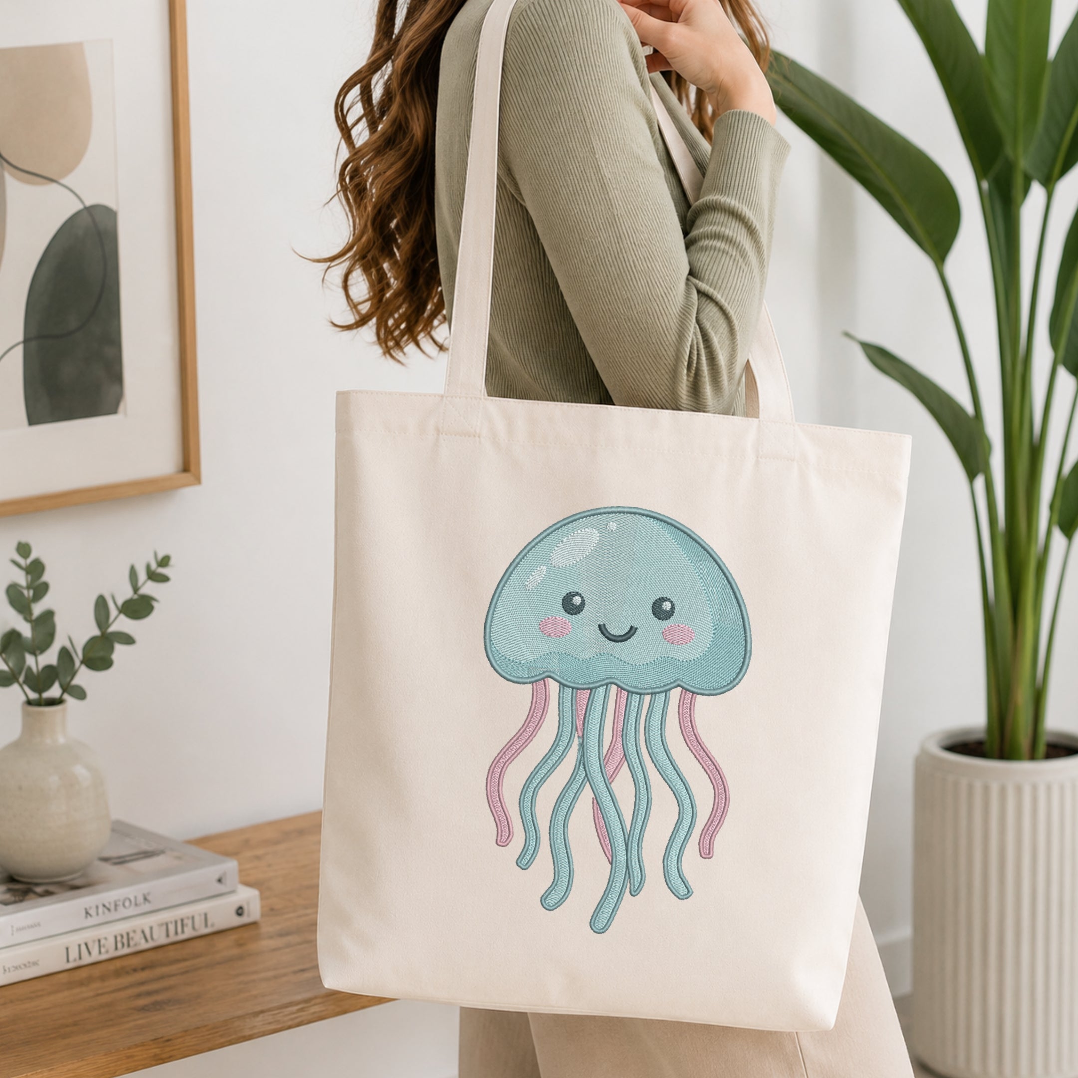 A woman wearing a sage green ribbed long-sleeve top carries a cream canvas tote bag with a cute light blue jellyfish graphic featuring pink tentacles, set in a bright, modern room with a wooden table and potted plants.