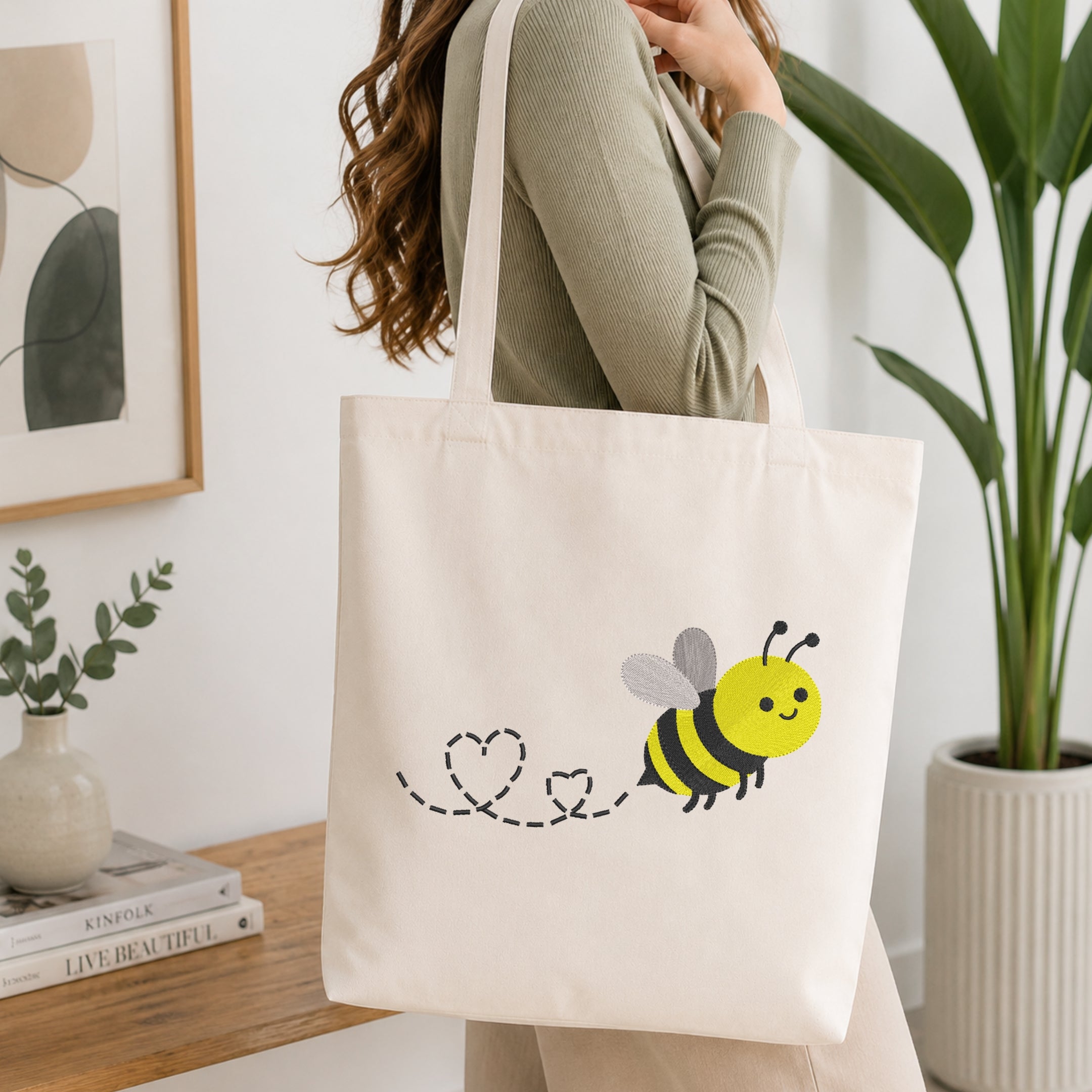 A woman wearing a sage green ribbed sweater carries a cream canvas tote bag with a cute yellow and black bumblebee graphic. The setting is a bright, modern room with a wooden shelf, a potted green plant, and a framed abstract print.