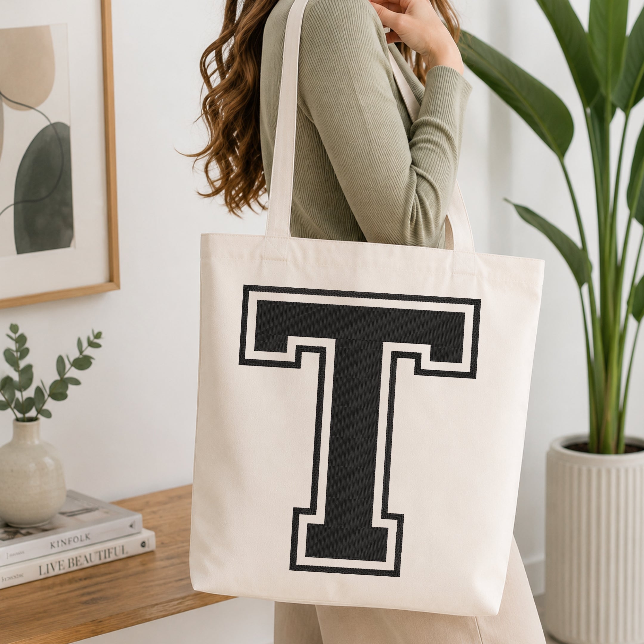 A person carrying a white canvas tote bag featuring a large embroidered varsity block letter 'T' in black with a white outline, set in a bright room with green house plants.