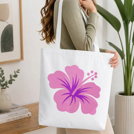 A woman wearing a sage green ribbed long-sleeve top carries a white canvas tote bag featuring a large pink and purple hibiscus flower graphic. The setting is a bright, modern room with a wooden table, books, a framed print, and a potted plant.