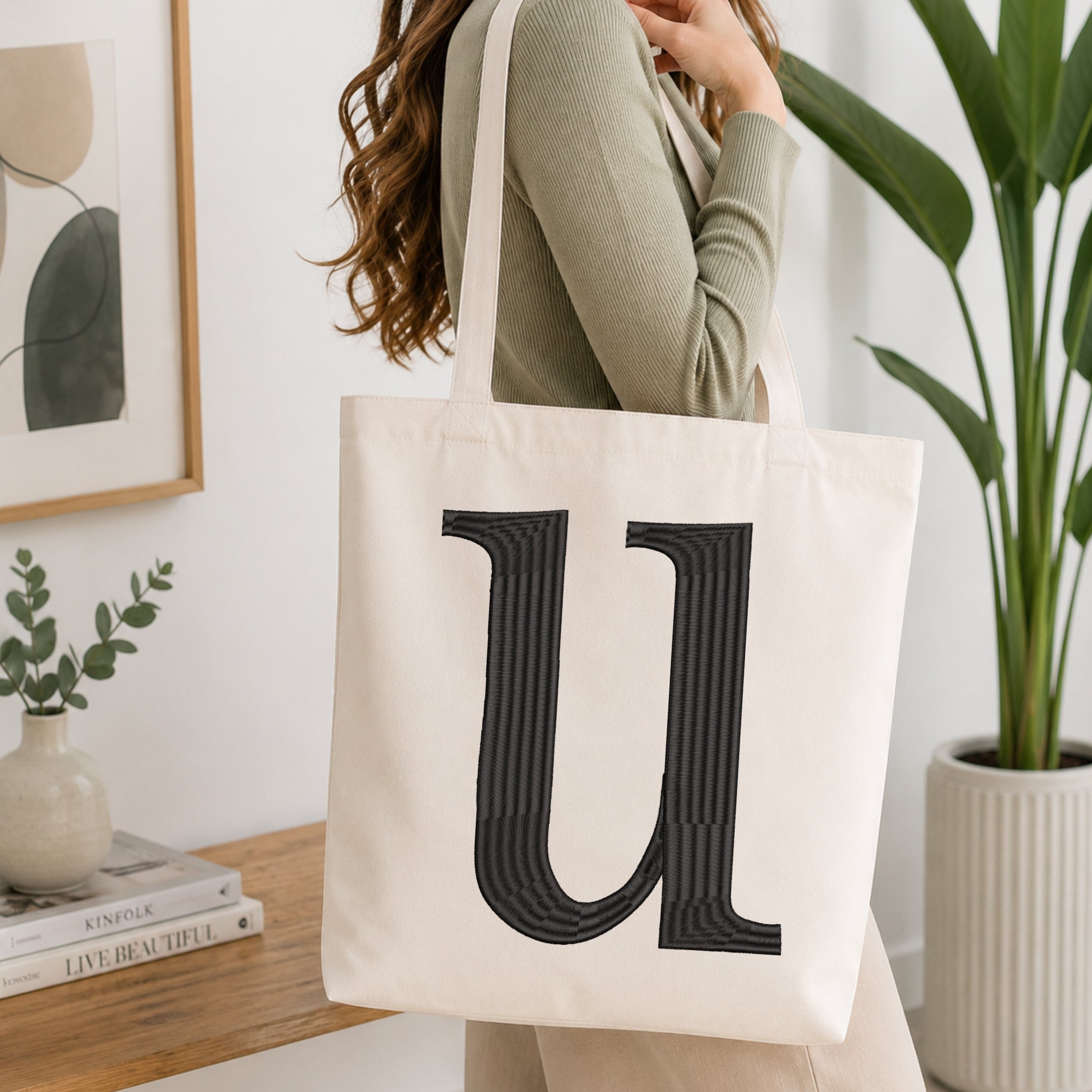 A woman wearing a sage green ribbed long-sleeve top carries a large off-white canvas tote bag with a bold black striped letter 'U' printed on the front, set in a bright, modern room with a wooden console table and a potted plant.